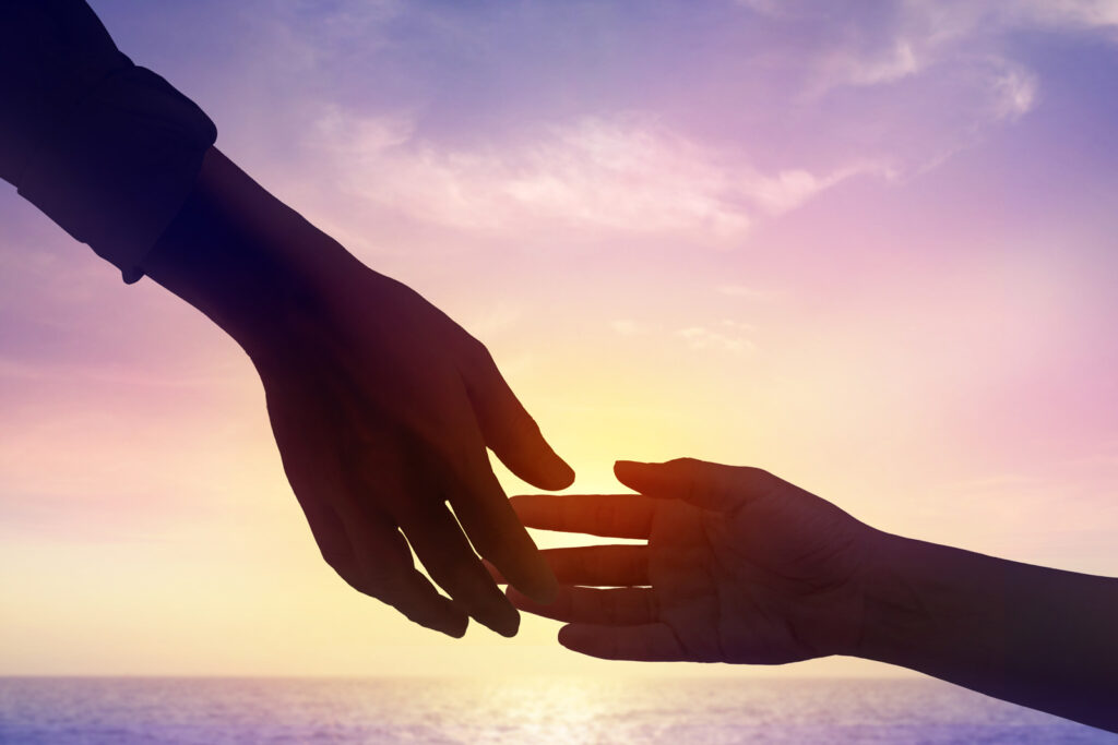 A close up of 2 different people's hands reaching for each other and just starting to connect. A sunset over the ocean is in the background.